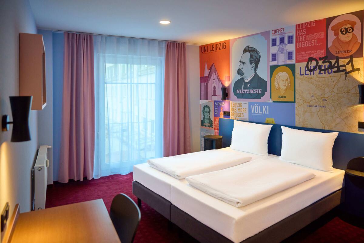 Habitación de hotel con cama doble, escritorio y silla. Hay carteles en la pared y una ventana con cortinas.