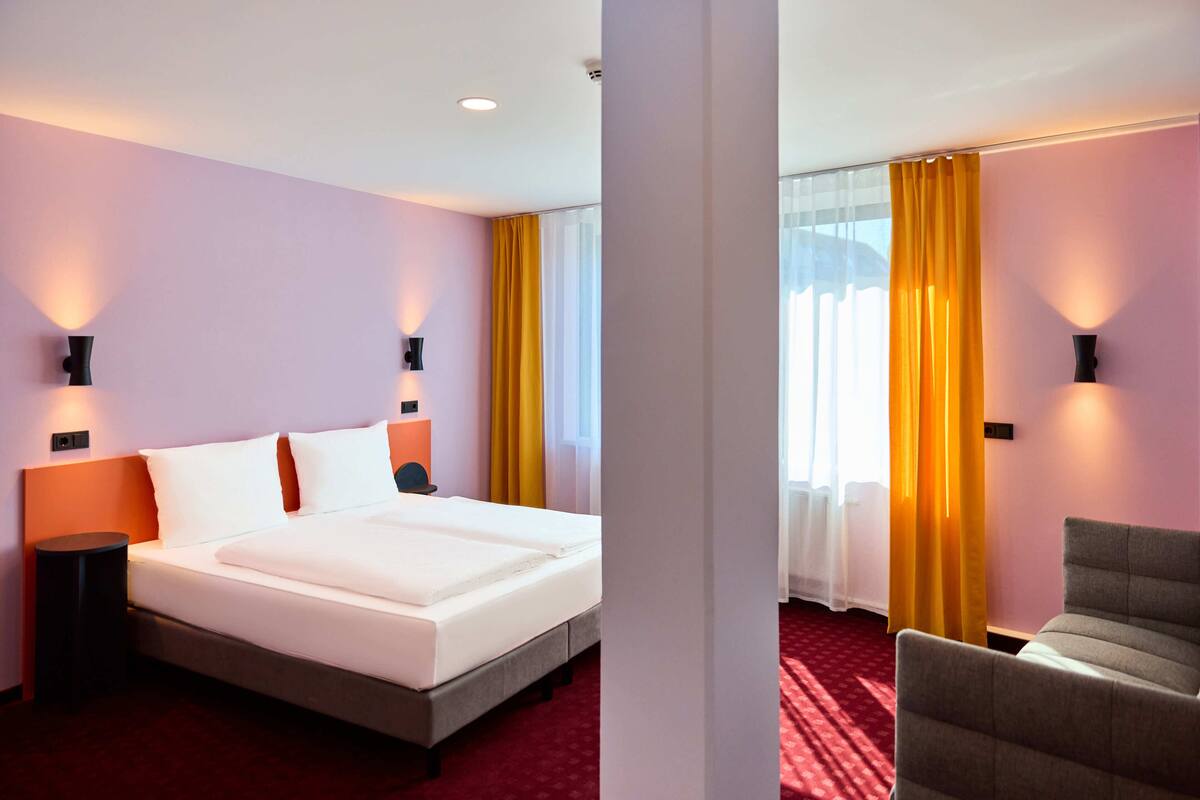 Habitación de hotel con una cama grande, dos mesitas de noche con lámparas, una silla y vistas al exterior a través de una ventana con cortinas amarillas.