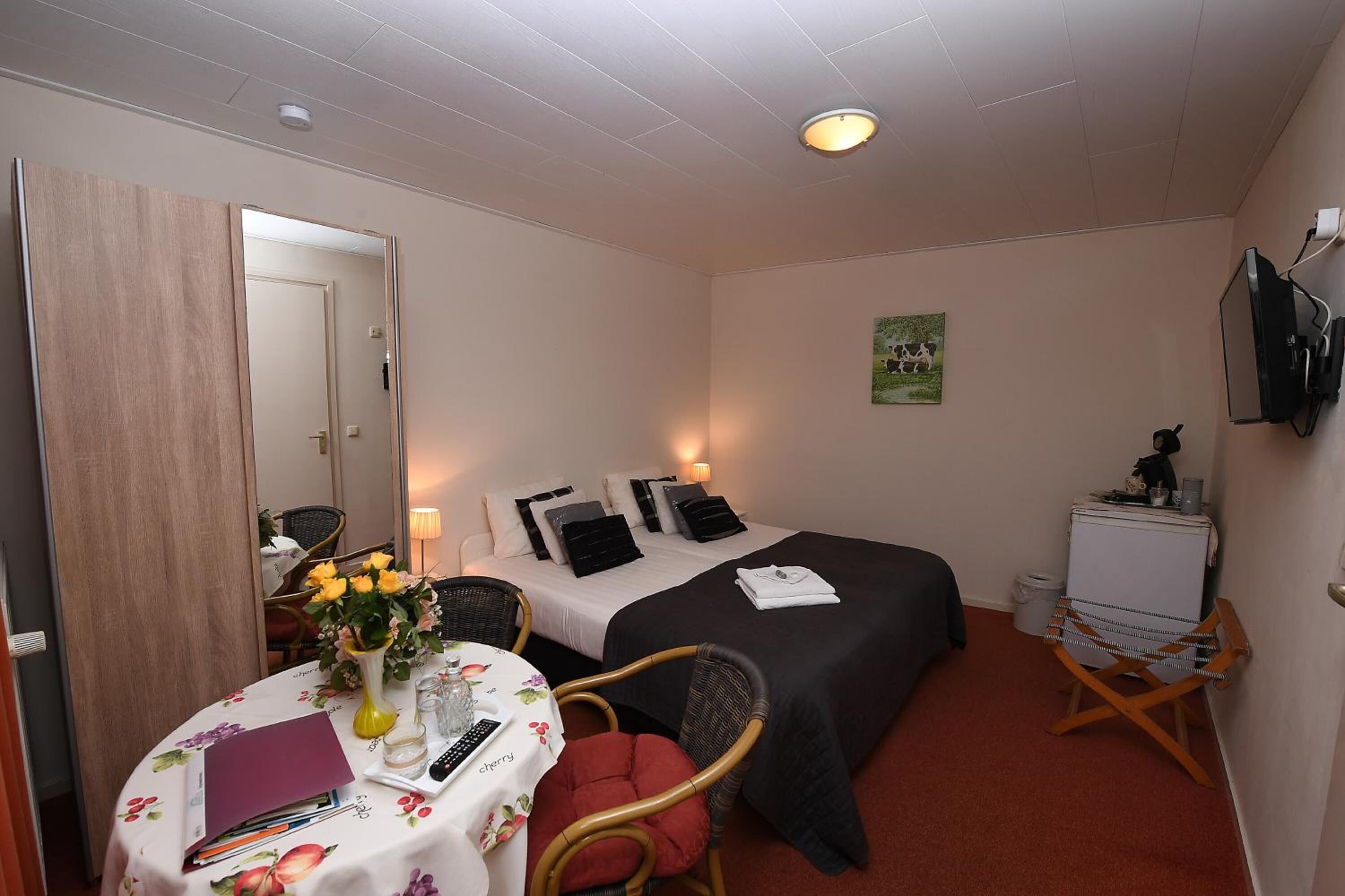 A hotel room with a bed, a desk with a chair, a small table with a vase of flowers, and a television on the wall.