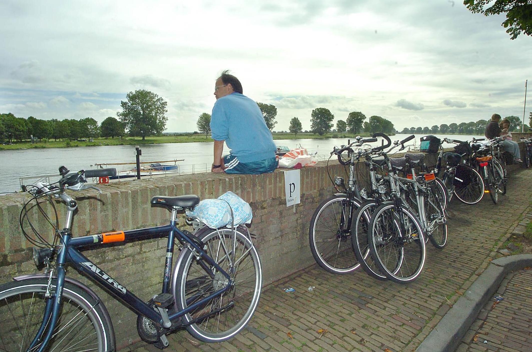 A man sitting on a wall overlooking a river with bicycles parked nearby.