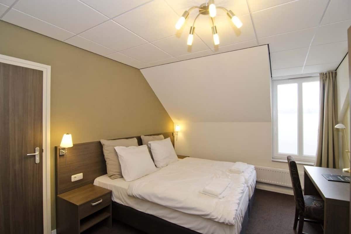 A hotel room with a bed, bedside table, lamp, desk, chair, and a window with curtains.