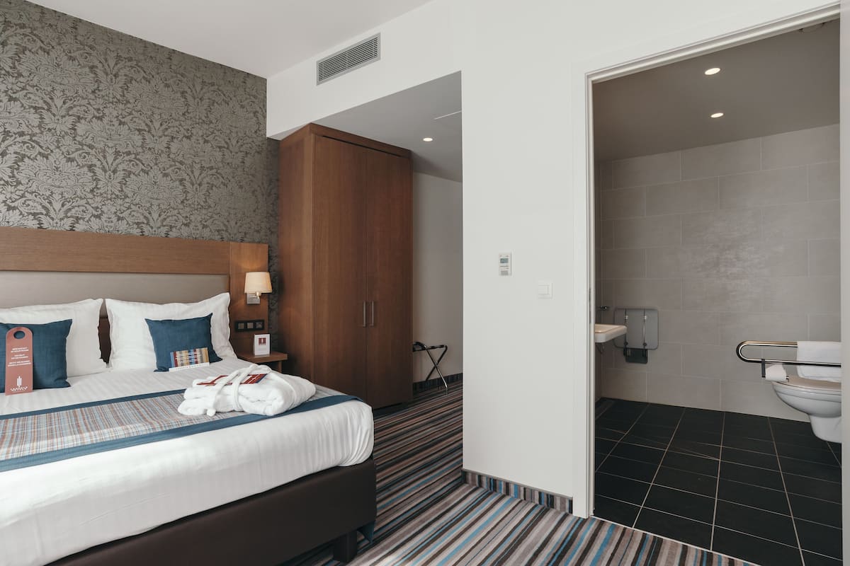 A hotel room with a large bed, a wooden wardrobe, a bathroom with a shower, and a patterned carpet.