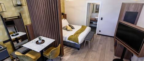 A modern hotel room with a bed, bedside table, and a TV. There is a dining area with a table and chairs.