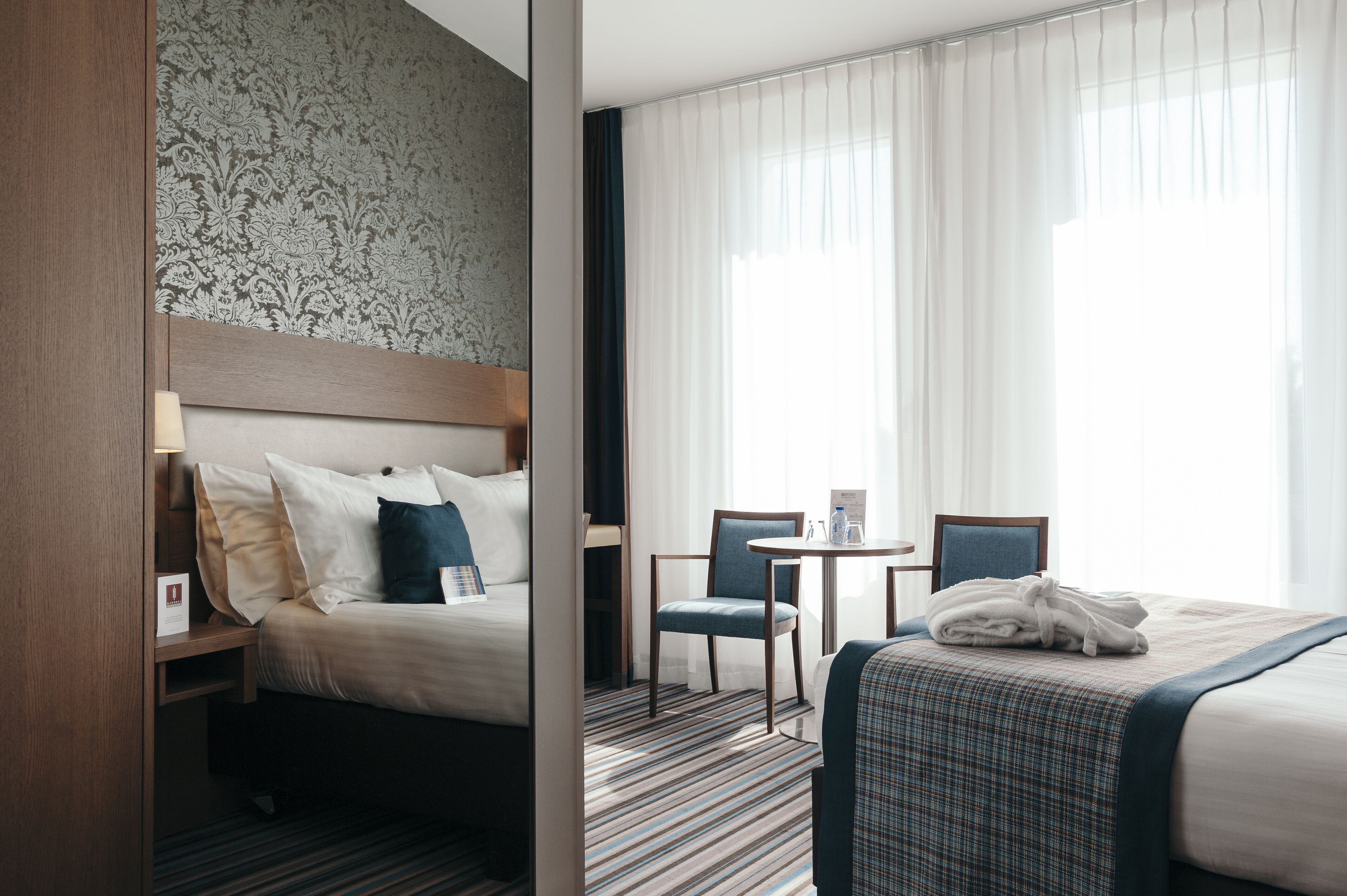 Une chambre d’hôtel avec un lit, une chaise, une petite table et une fenêtre avec des rideaux.