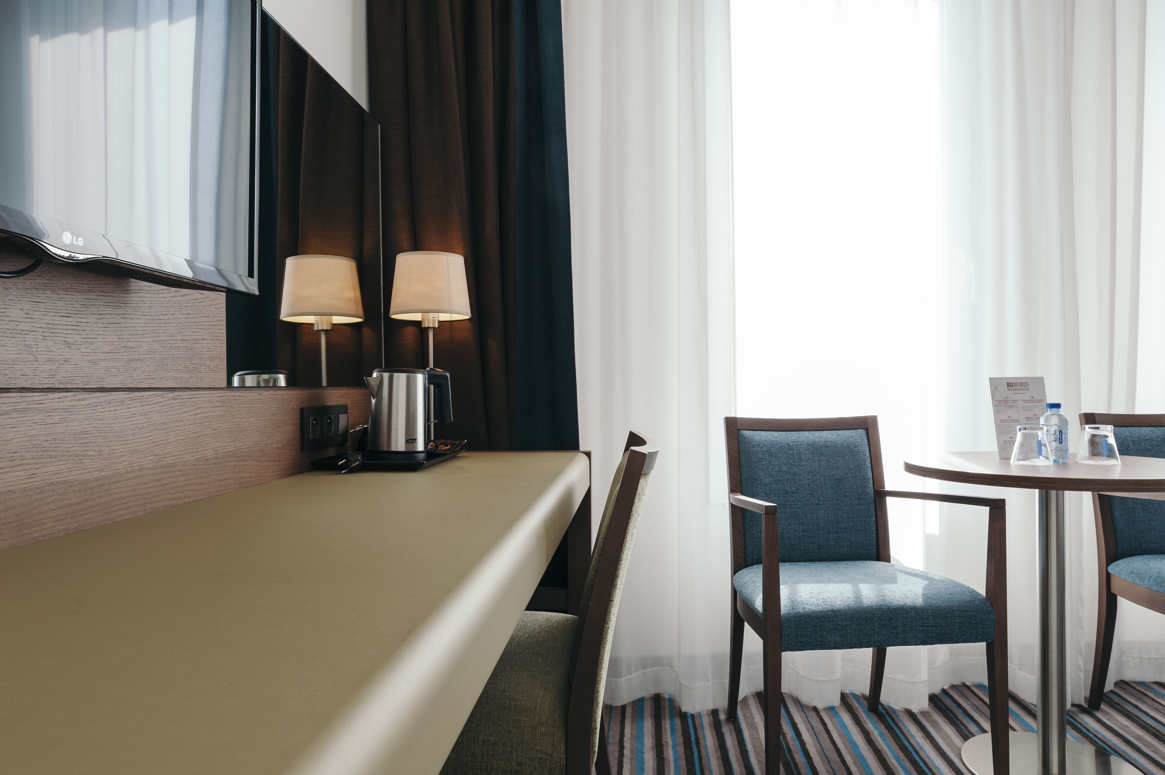 Habitación de hotel con una cama, un escritorio con dos lámparas, una silla, una mesita con una botella de agua y una ventana con cortinas.