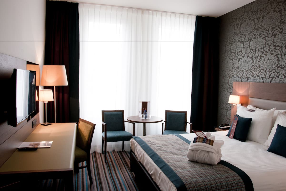 A hotel room with a bed, two chairs, a small table, a desk, a TV, and a window with curtains.