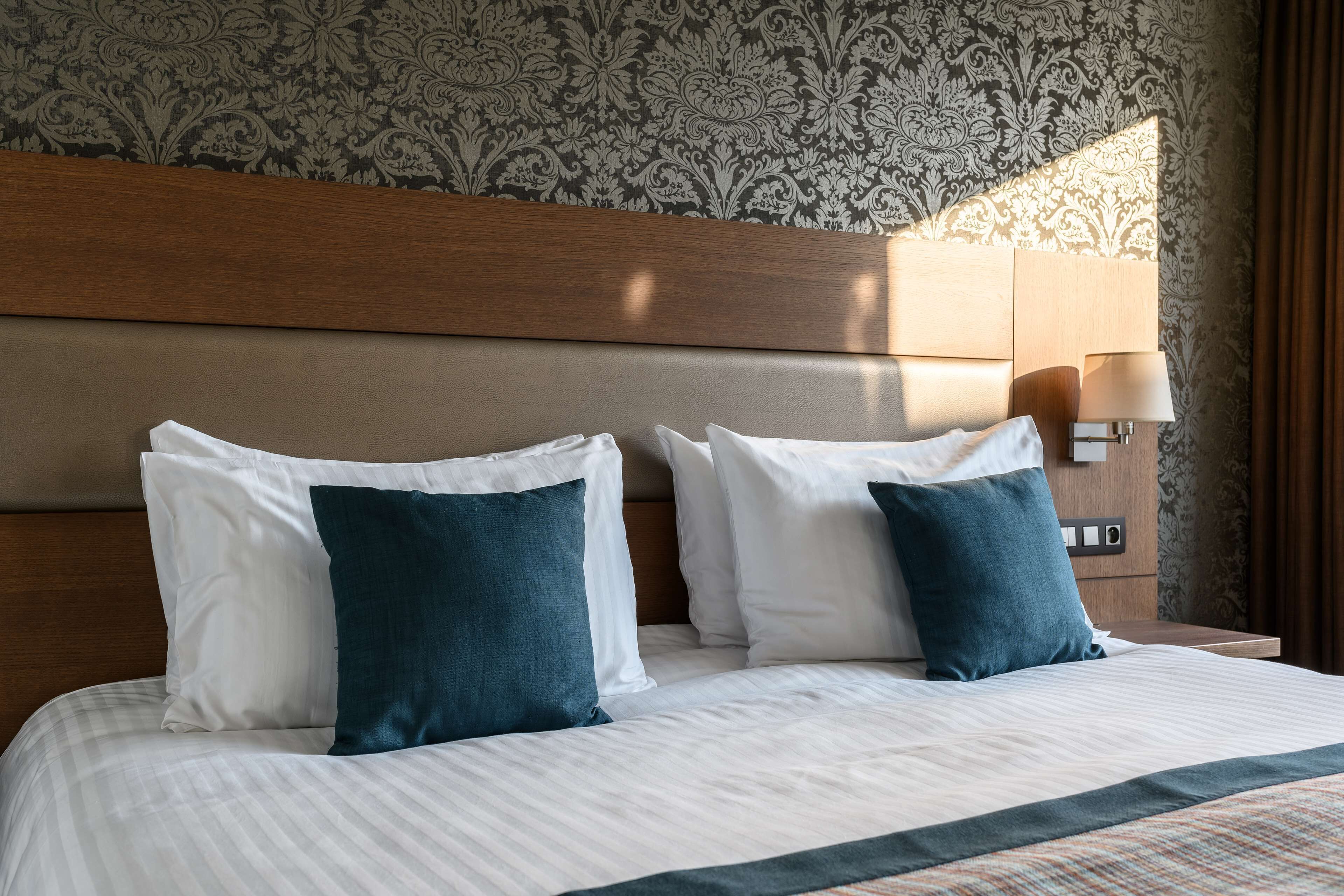Een net opgemaakt bed met witte en donkerblauwe kussens, een houten hoofdbord en behang met een patroon.