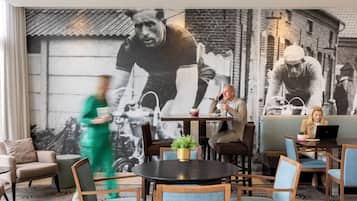 A modern hotel lobby with a large mural of cyclists, comfortable seating, and a coffee table with a plant.