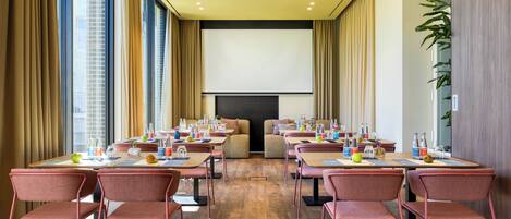 Un salón de conferencias con mesas, sillas, una pantalla de proyector y un amplio ventanal con cortinas.