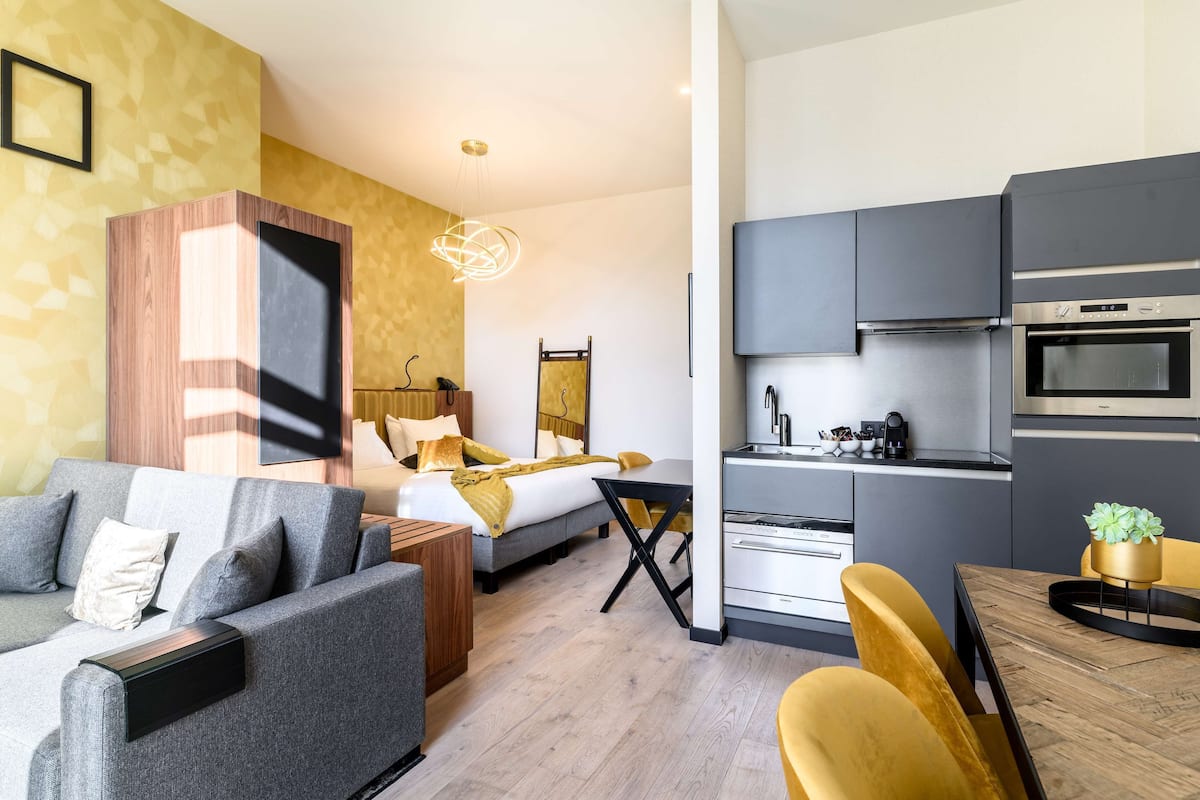 A modern hotel room with a sofa, a bed, a dining table with chairs, a kitchen area with a stove and oven, and a pendant light.
