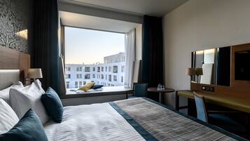 A hotel room with a large bed, a desk, a chair, a TV, and a window with a view of buildings.