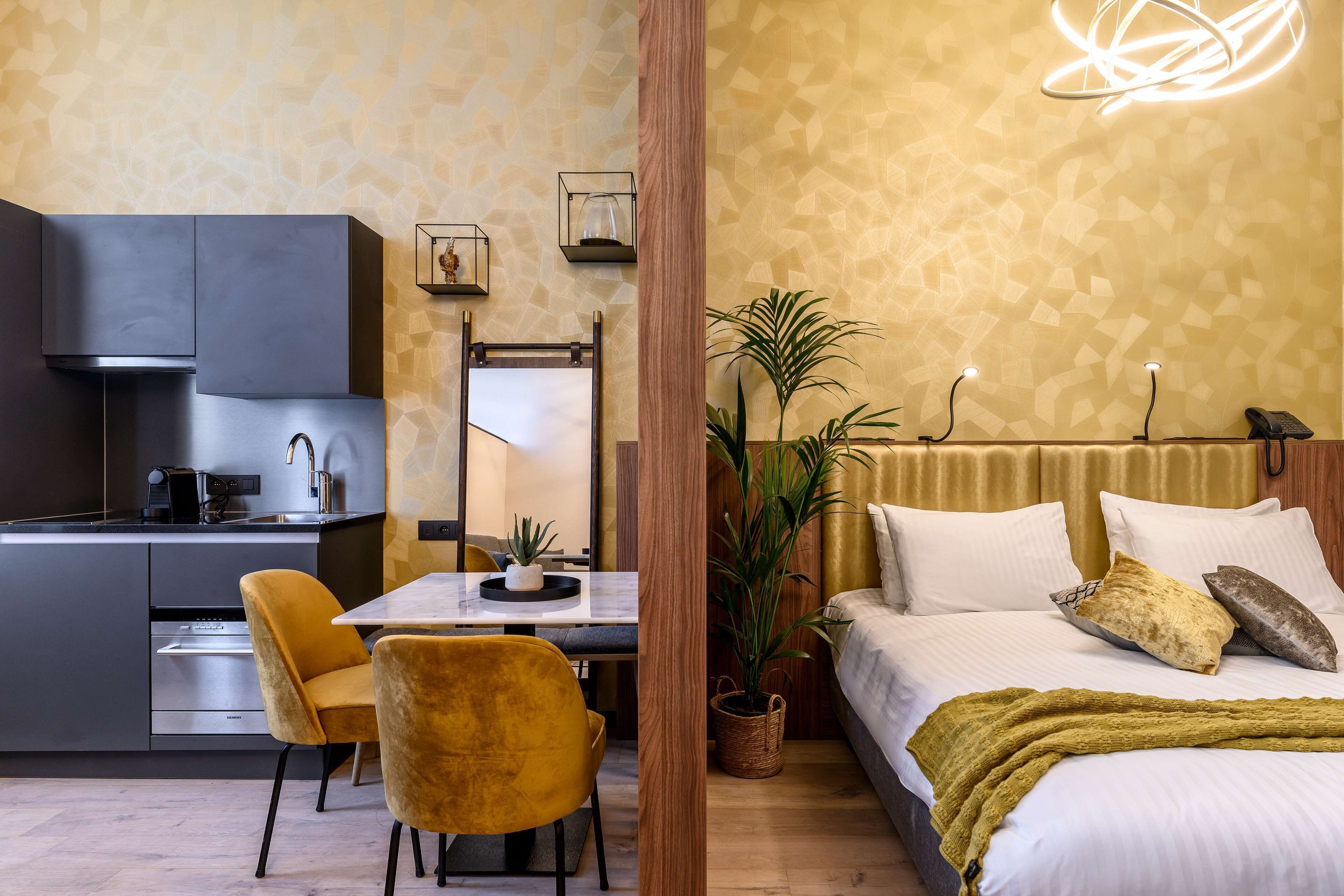 A modern hotel room with a kitchen, dining area, and a large bed with a green throw blanket.