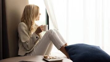 A person sitting by a window with a cup, a remote control, and a plate with food on a small table.