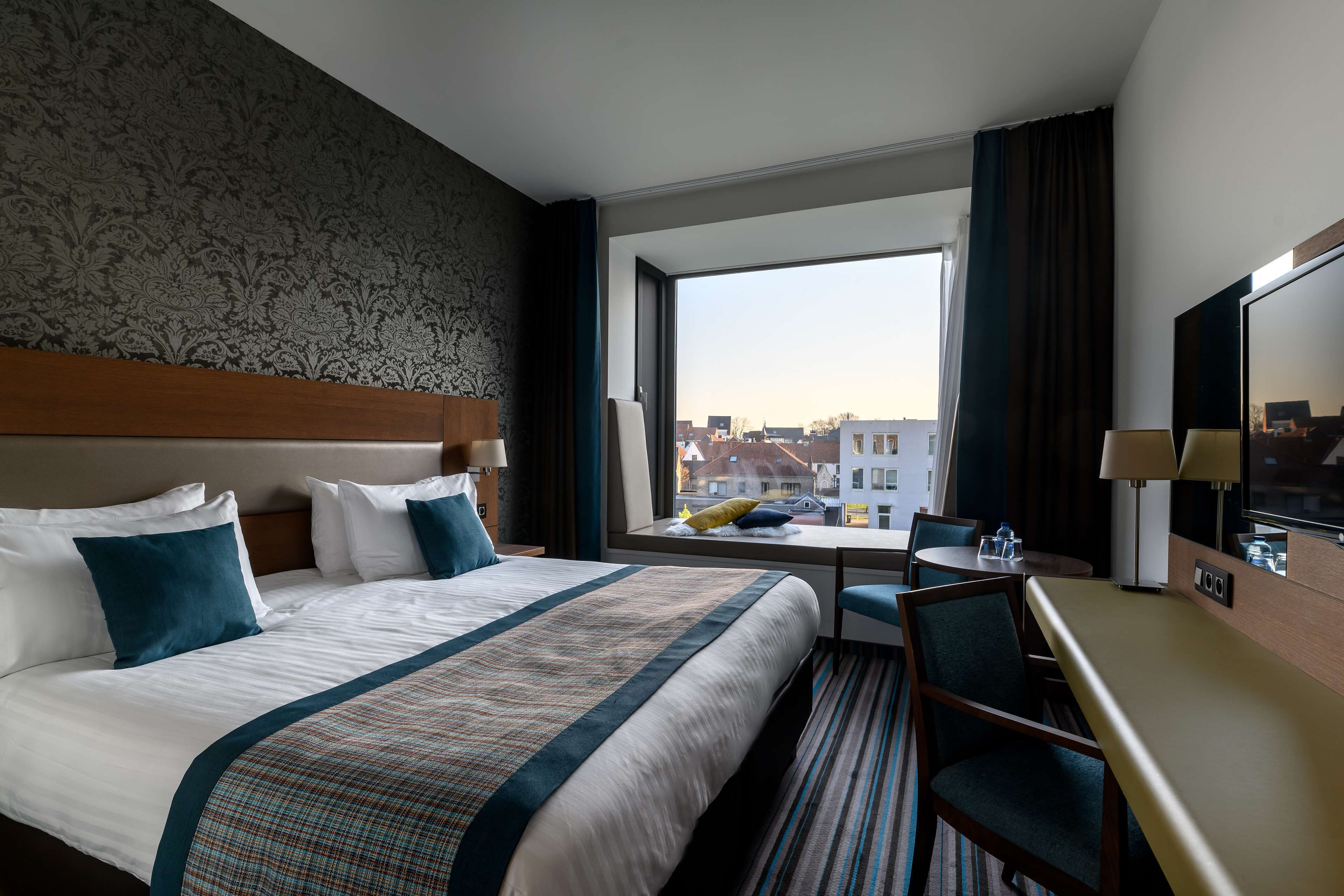 A hotel room with a large bed, a desk, and a view of buildings through a window.