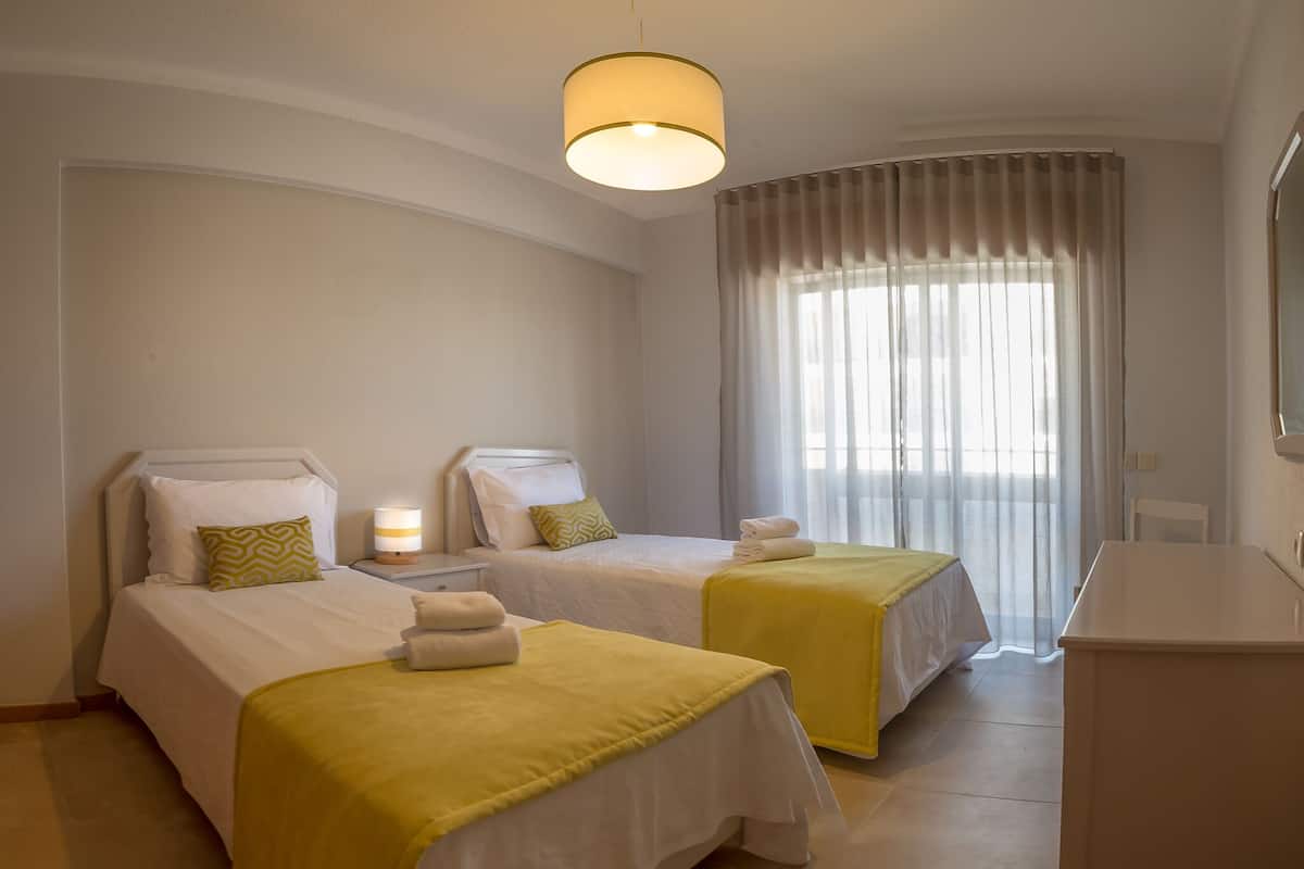 A hotel room with two beds, a desk, a lamp, and a large window with curtains.