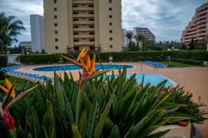A hotel pool area with a large building in the background, surrounded by greenery and blue lounge chairs.