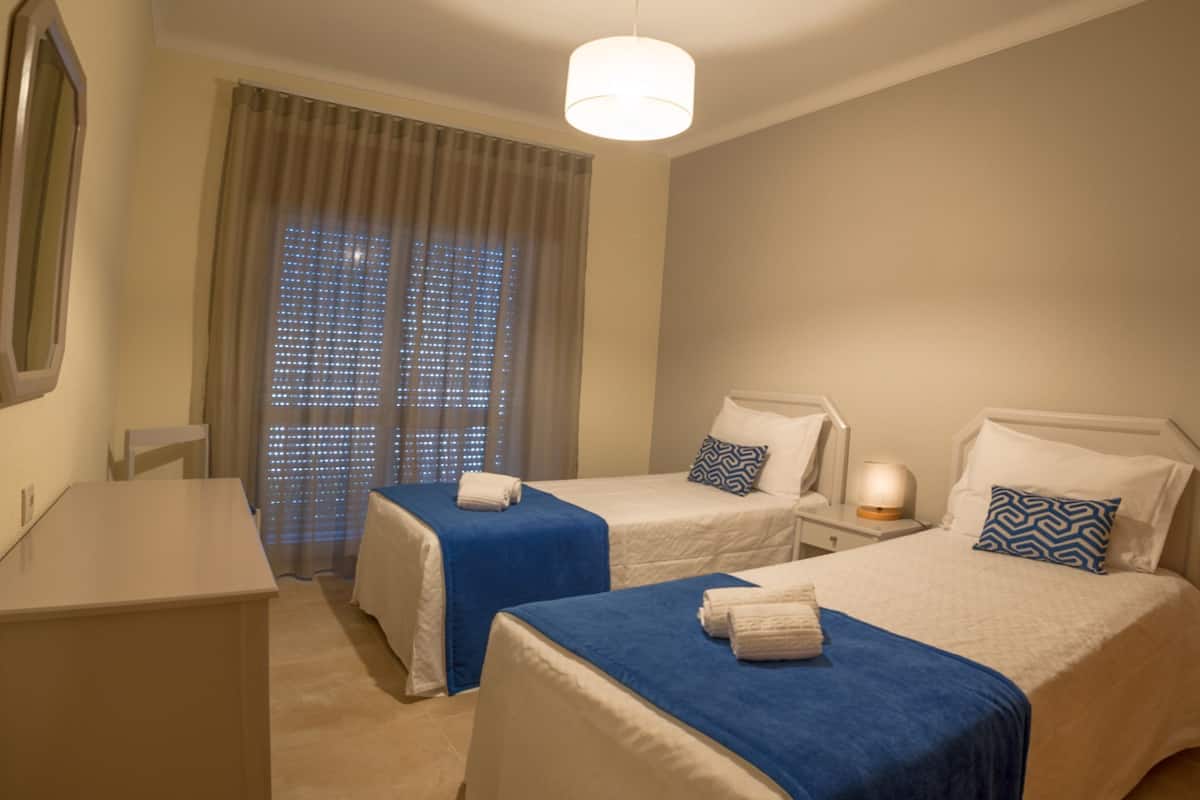 A hotel room with two beds, a desk, a mirror, a window with curtains, and a hanging light fixture.