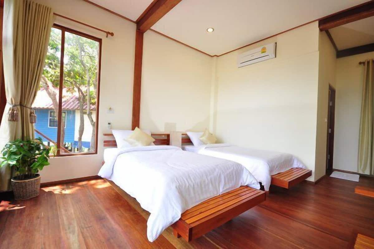 A room with two beds, wooden flooring, a potted plant, and a window with curtains.