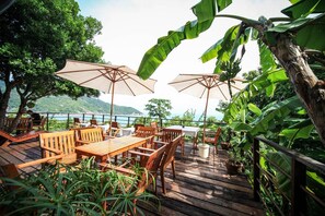 An outdoor patio with wooden tables and chairs, large umbrellas, and lush greenery.