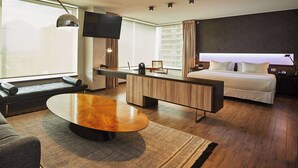 Moderni hotellihuone, jossa on suuri sänky, työpöytä tuolilla, sohva, sohvapöytä ja seinälle asennettu televisio.