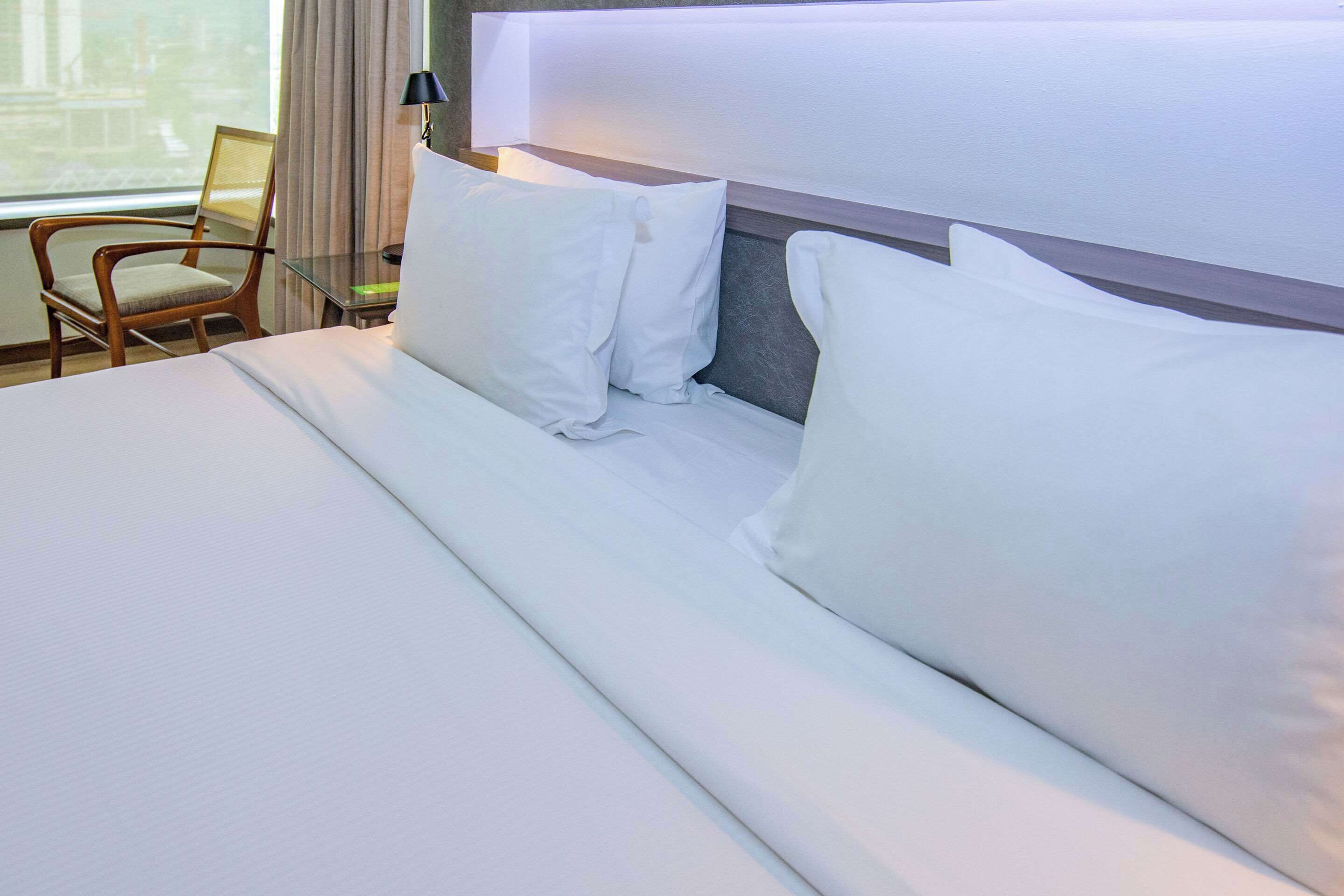 A neatly made bed with white linens and pillows, a wooden chair, and a bedside table with a lamp.