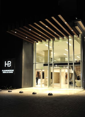Uma entrada de hotel moderna com portas de vidro, uma área de recepção visível e uma escadaria que leva a um andar superior.
