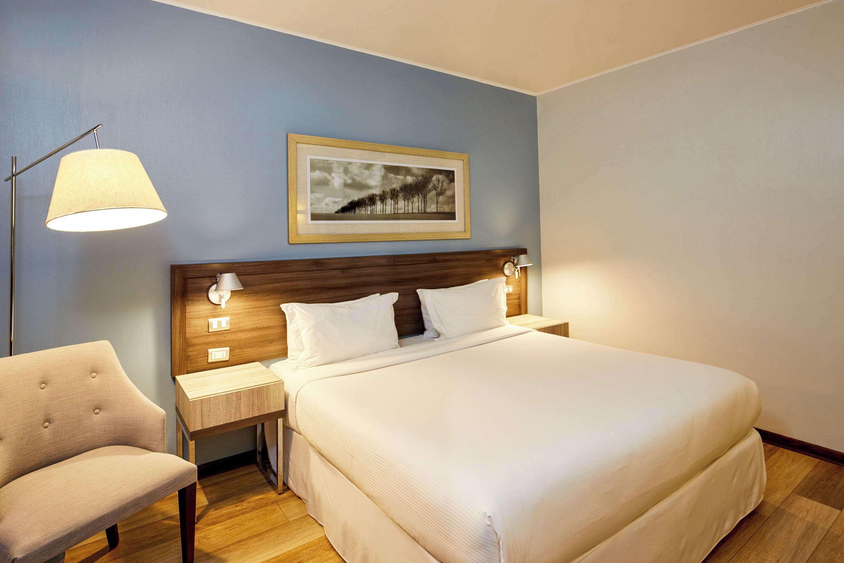 A hotel room with a large bed, a nightstand, a chair, a framed picture on the wall, and a floor lamp.