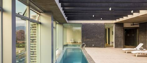 Una piscina interior moderna con paredes de vidrio transparente, techo de hormigón y vistas a la ciudad.