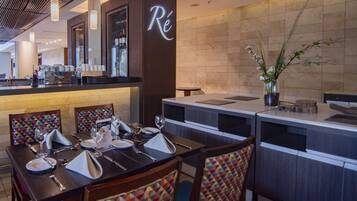 A restaurant with a modern interior, featuring a bar area with a wine cooler and a counter with a 'Ré' sign. There are tables set with wine glasses, plates, and cutlery.