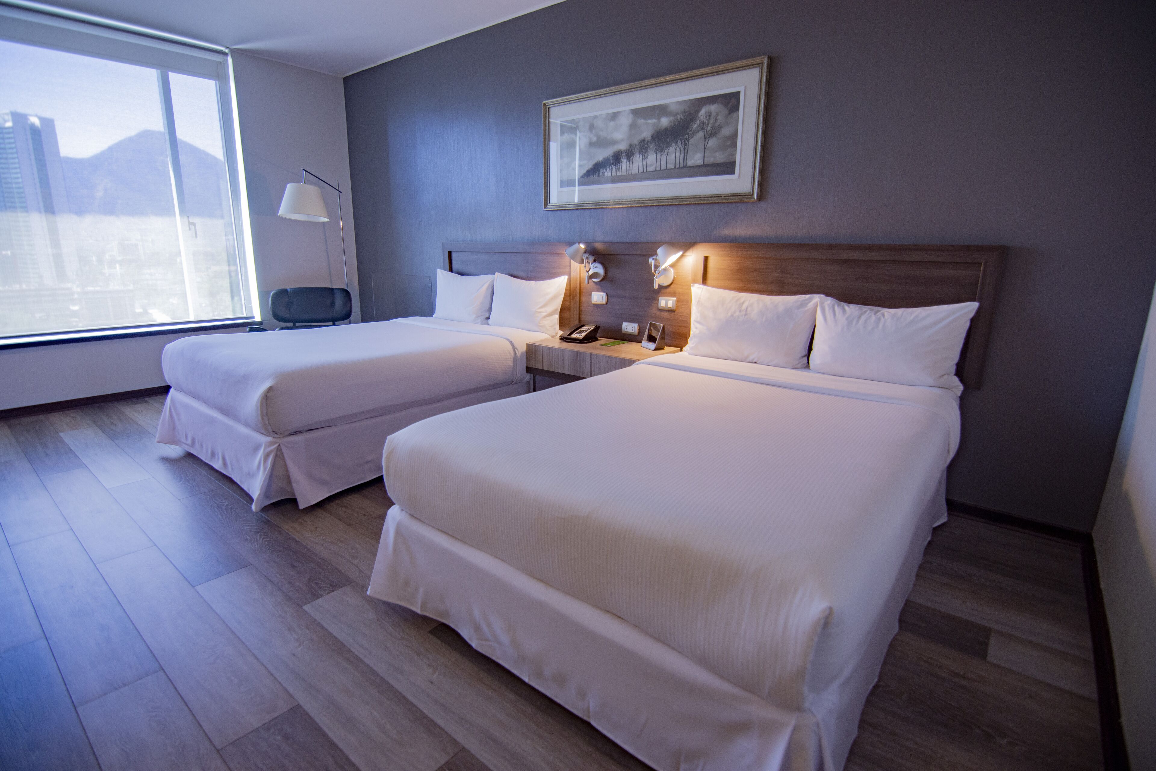 Habitación de hotel con dos camas, un ventanal grande, un cuadro en la pared y una mesita de noche con lámparas.