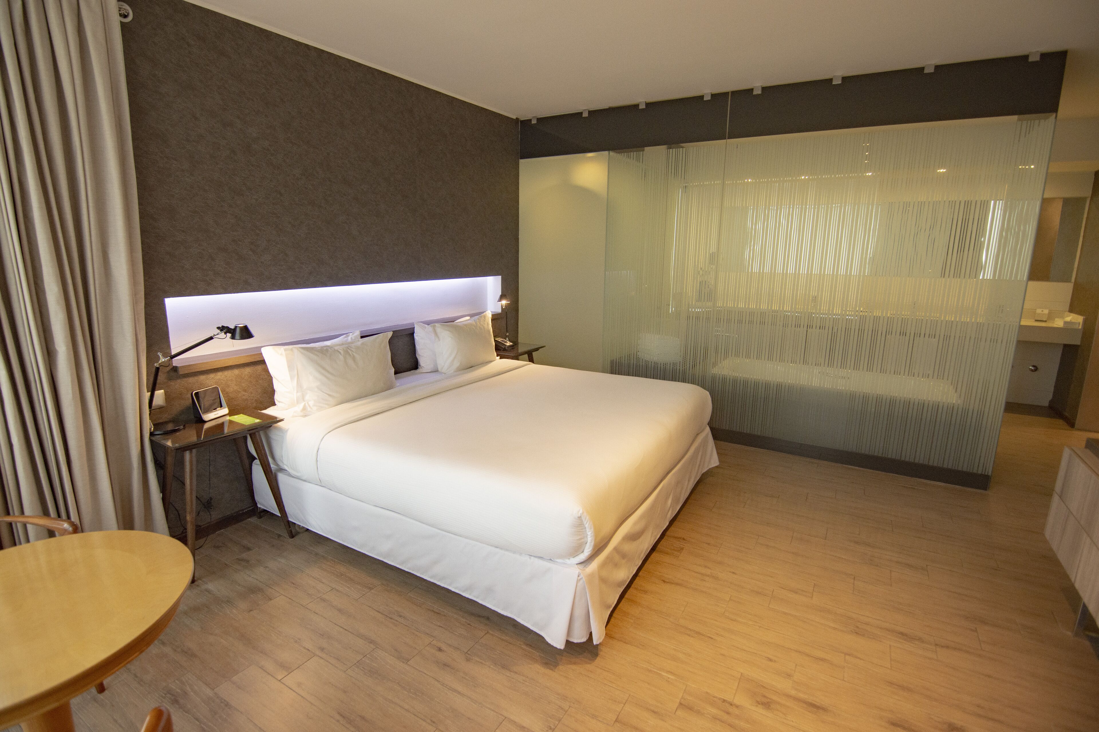 Moderni hotellihuone, jossa on suuri sänky, yöpöytä lampun kanssa, pieni työpöytä ja lasinen väliseinä.