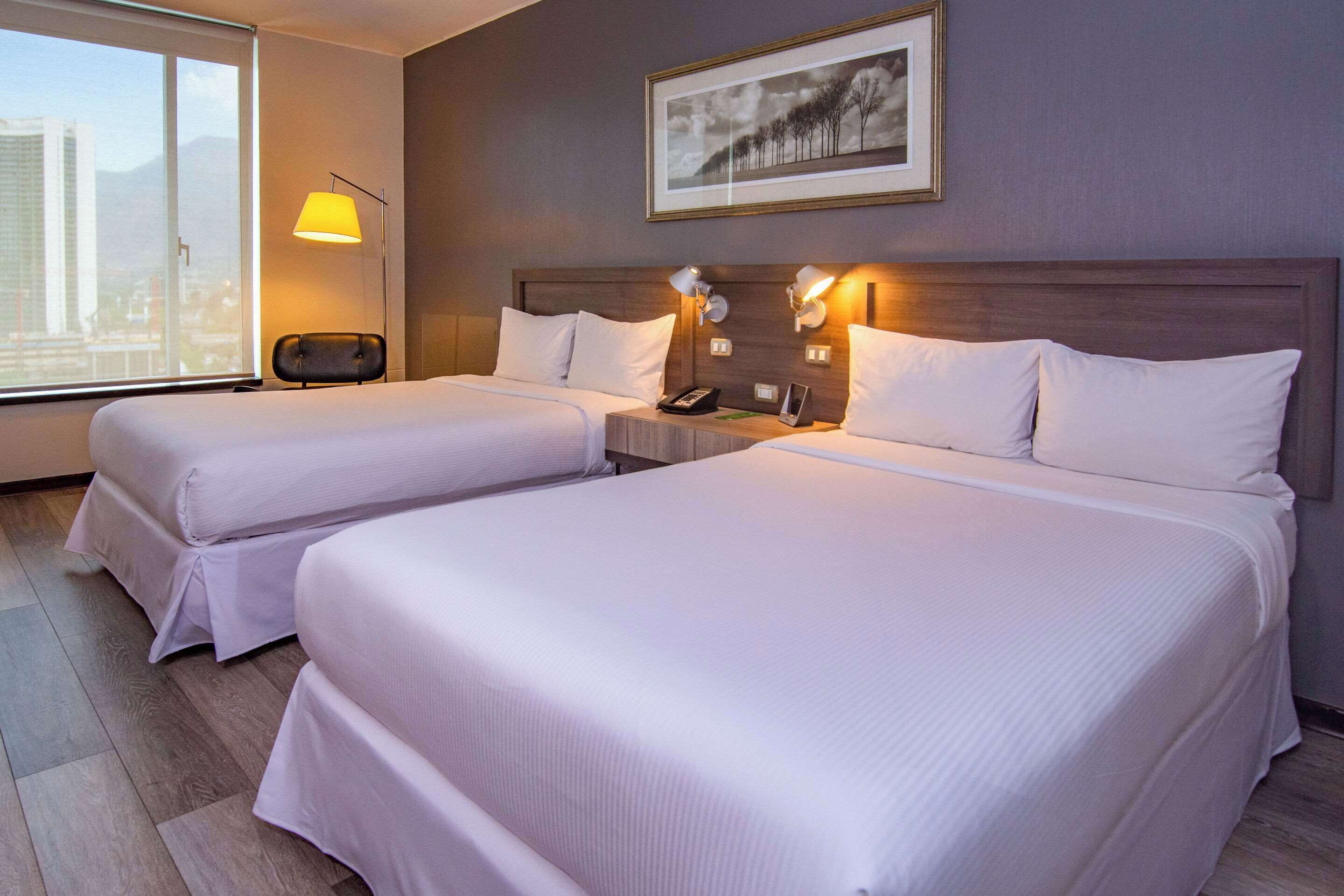 A hotel room with two beds, a nightstand, a lamp, a framed picture on the wall, and a window with a city view.
