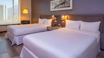 A hotel room with two beds, a nightstand, a lamp, a framed picture on the wall, and a window with a city view.