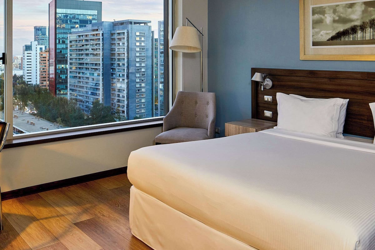 Une chambre d’hôtel avec un grand lit, une chaise, une table de chevet, une lampe et une fenêtre donnant sur la ville.