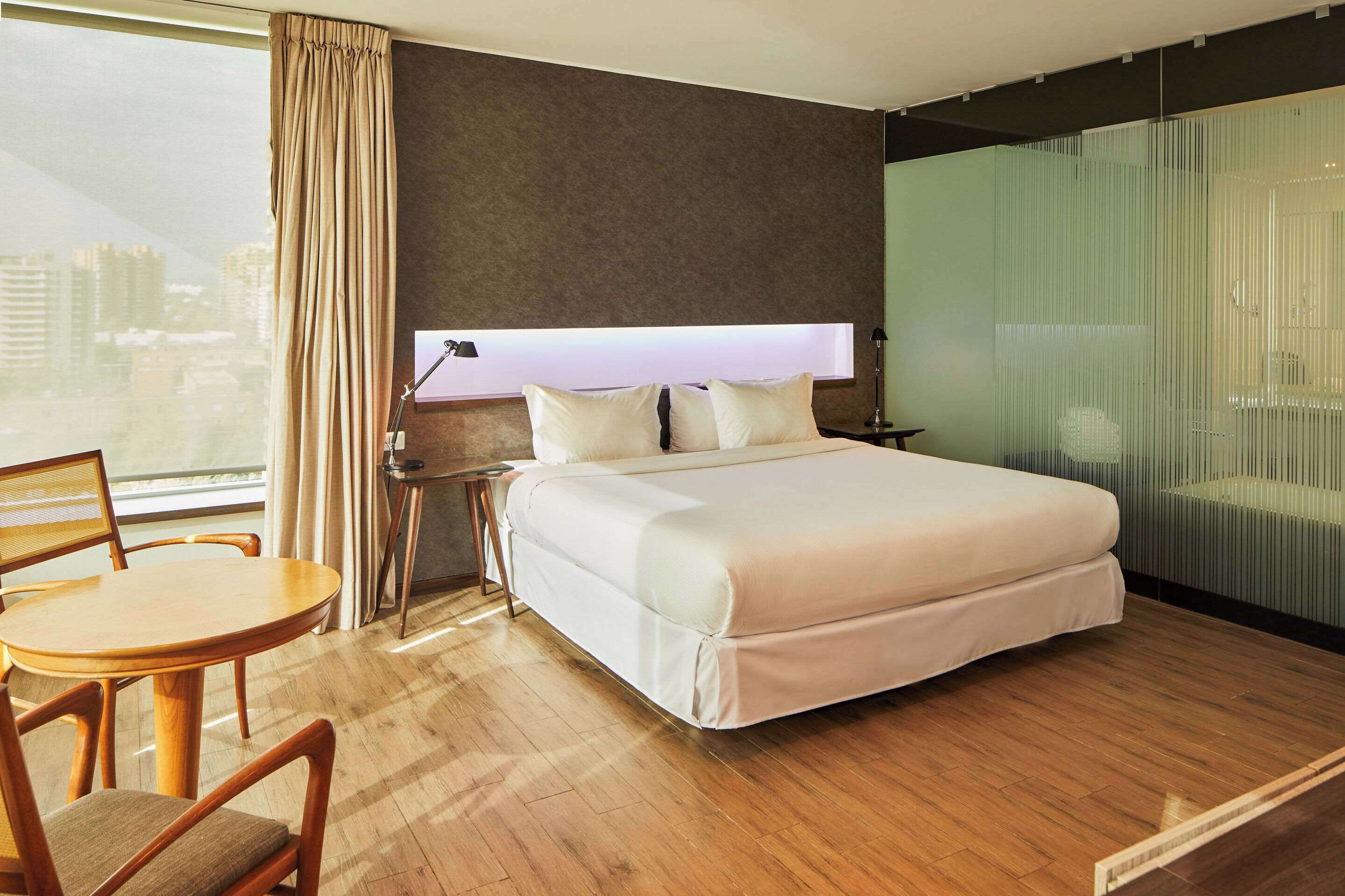 Ein modernes Hotelzimmer mit einem großen Bett, einem Schreibtisch, einem Sessel und Blick auf die Stadt.