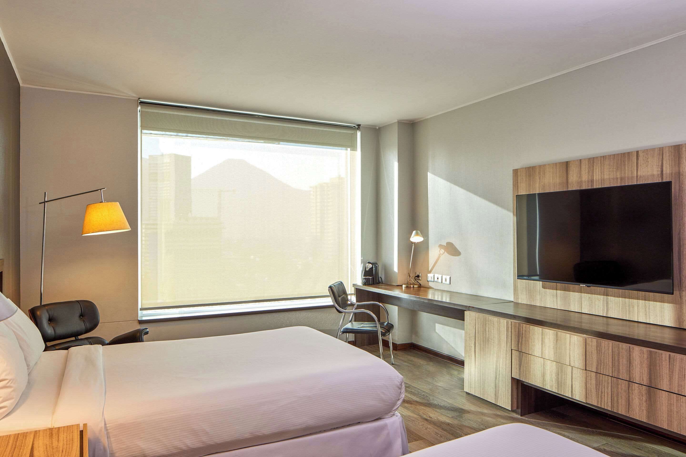 A hotel room with two beds, a desk, a chair, a TV, and a large window.