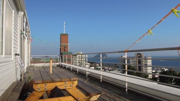 Un balcón con vista al mar y edificios, una banca de madera y un tendedero con ropa colgada.