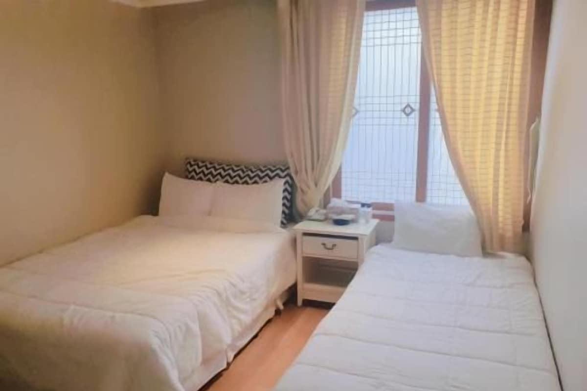 A small, single-room hotel room with two beds, a nightstand, and a window with curtains.