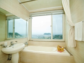 A bathroom with a bathtub, a sink, and a window offering a city view. - Good Inn Hotel (Seogwipo)