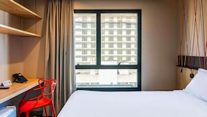 A hotel room with a bed, a desk with a telephone, a chair, and a window with a view of buildings. - Ibis Styles Porto Alegre Moinhos de Vento (Porto Alegre)