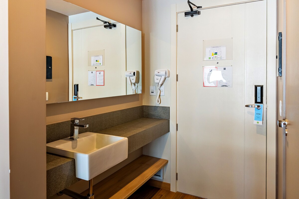 Een badkamer met een wastafel, spiegel, handdoekhouder en een deur met bedieningspaneel.
