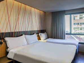 Een hotelkamer met twee bedden, een modern hoofdeinde, een raam met gordijnen en uitzicht op gebouwen.