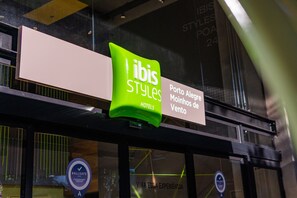 Letrero de 'ibis Styles Hotels' con un logo verde y texto que indica 'Porto Alegre' y 'Moninhos de Vento'.
