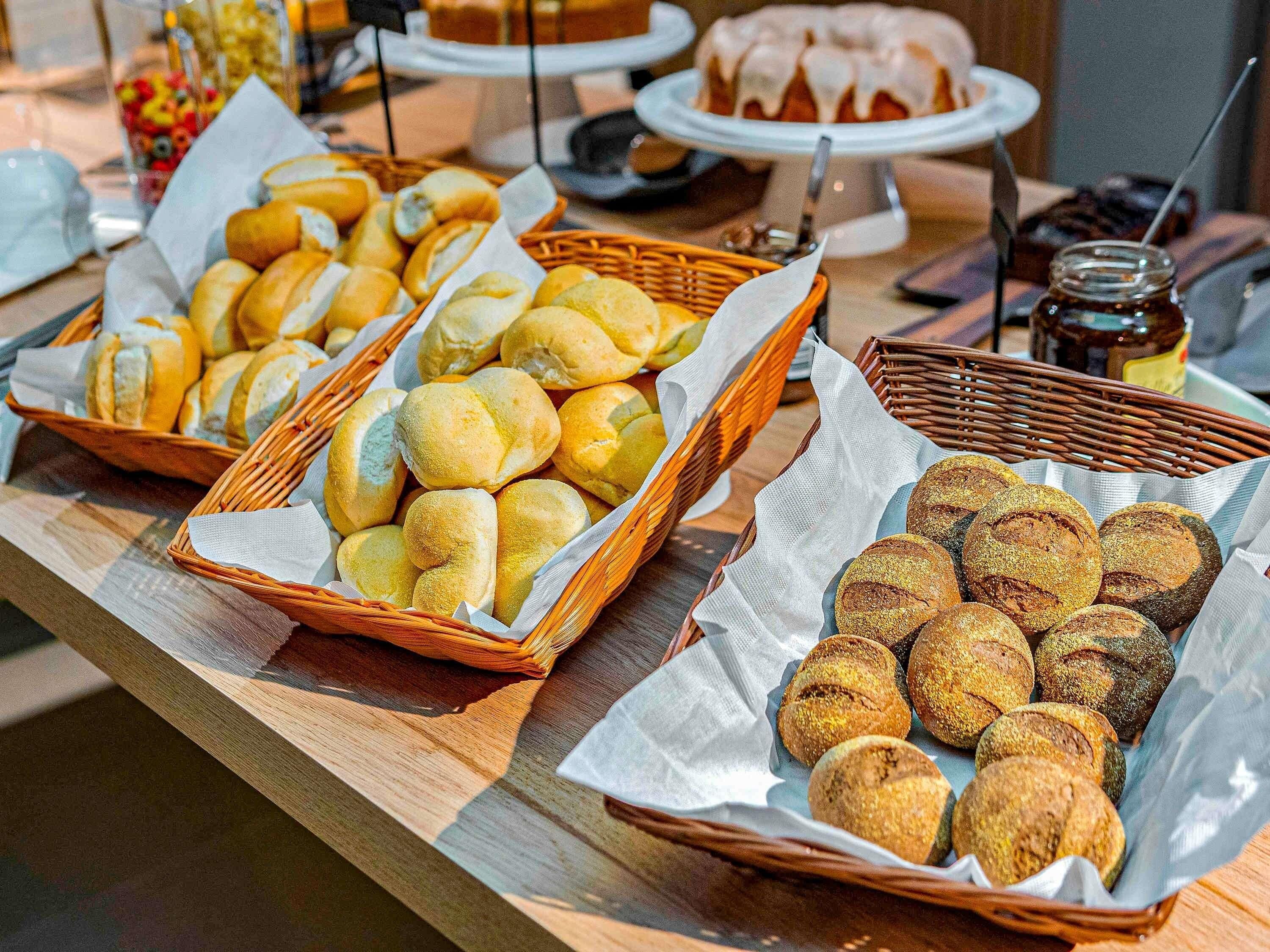 Uma mesa de buffet com diversos produtos de panificação, incluindo pães e doces, dispostos em cestos.