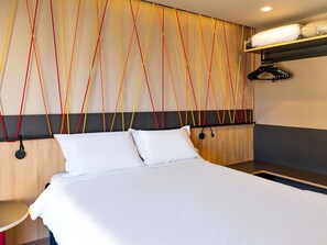 A hotel room with a bed, a bedside table, a wall with colorful lines, a coat rack, and a shelf with pillows. - Ibis Styles Porto Alegre Moinhos de Vento (Porto Alegre)