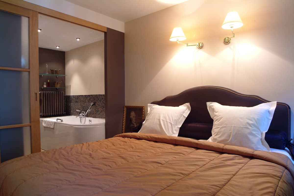 A neatly made bed with a brown comforter and white pillows, a dark headboard, and a wall-mounted light fixture.