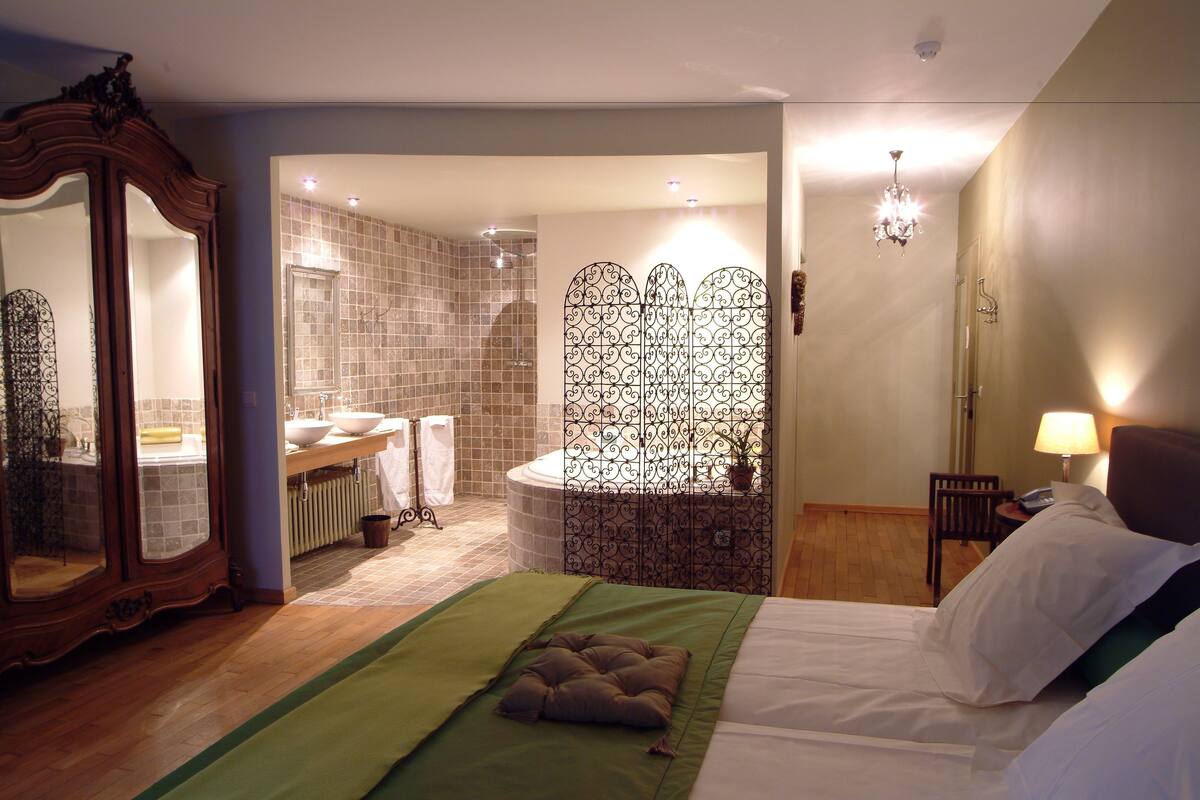 A hotel room with a bed, a bathroom with a bathtub, a mirror, and a chandelier.