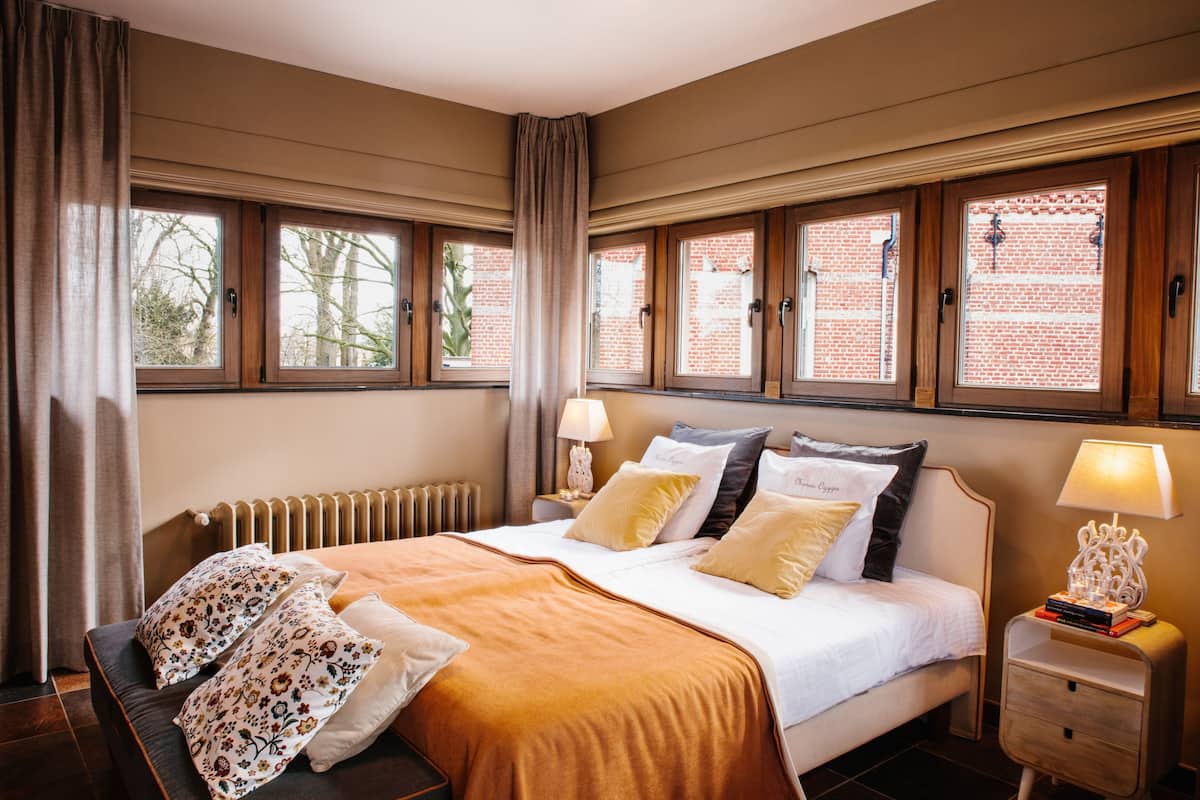 A hotel room with a large bed, a nightstand with a lamp, and a view of a brick building through the windows.