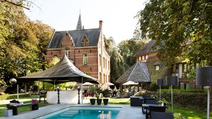 A swimming pool with outdoor seating and umbrellas, surrounded by trees and a large building with a tower. - Charmehotel Manoir Ogygia (Poperinge)