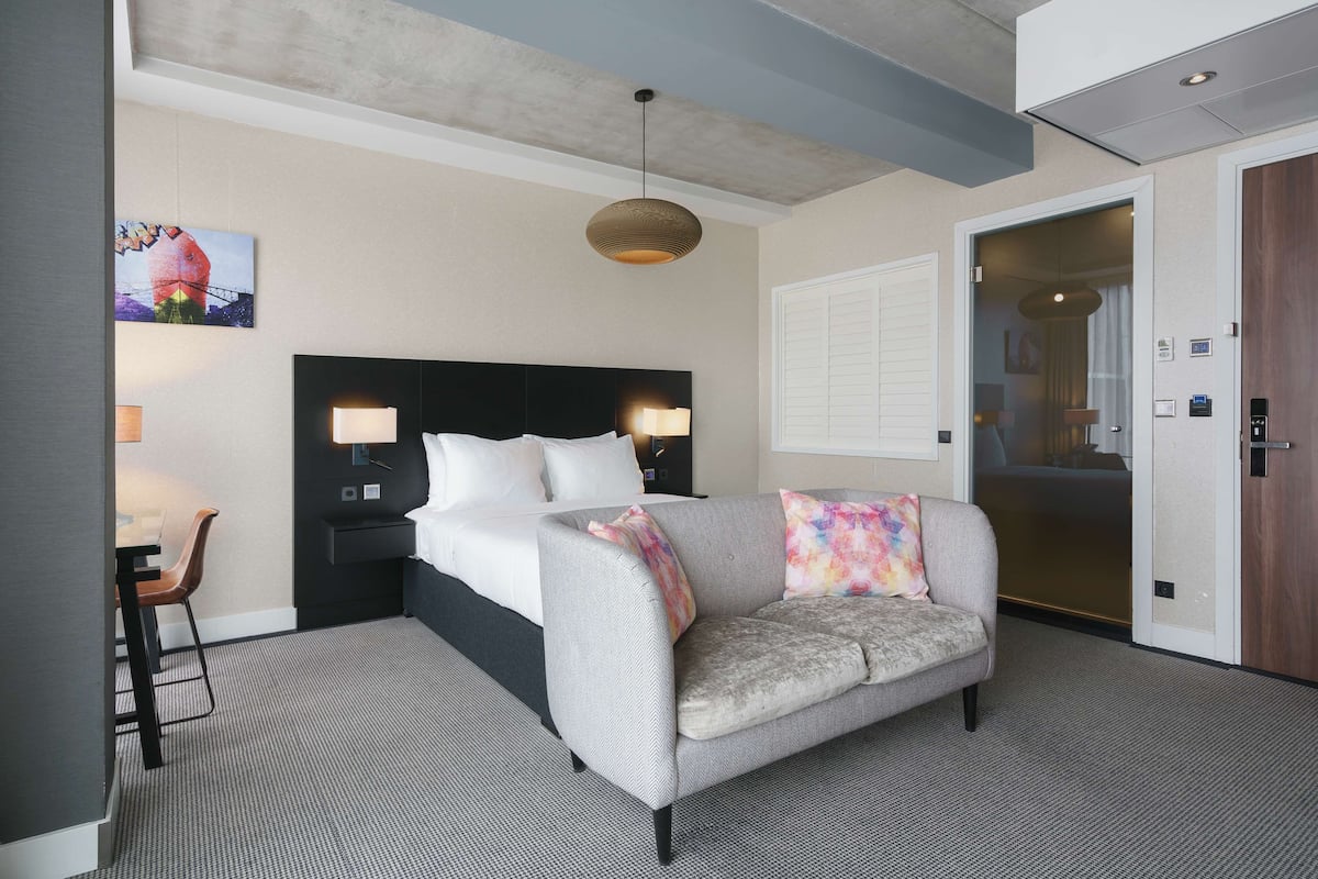Une chambre d’hôtel comprenant un lit, un canapé, un bureau avec une chaise, un luminaire suspendu et une porte donnant sur une autre pièce.